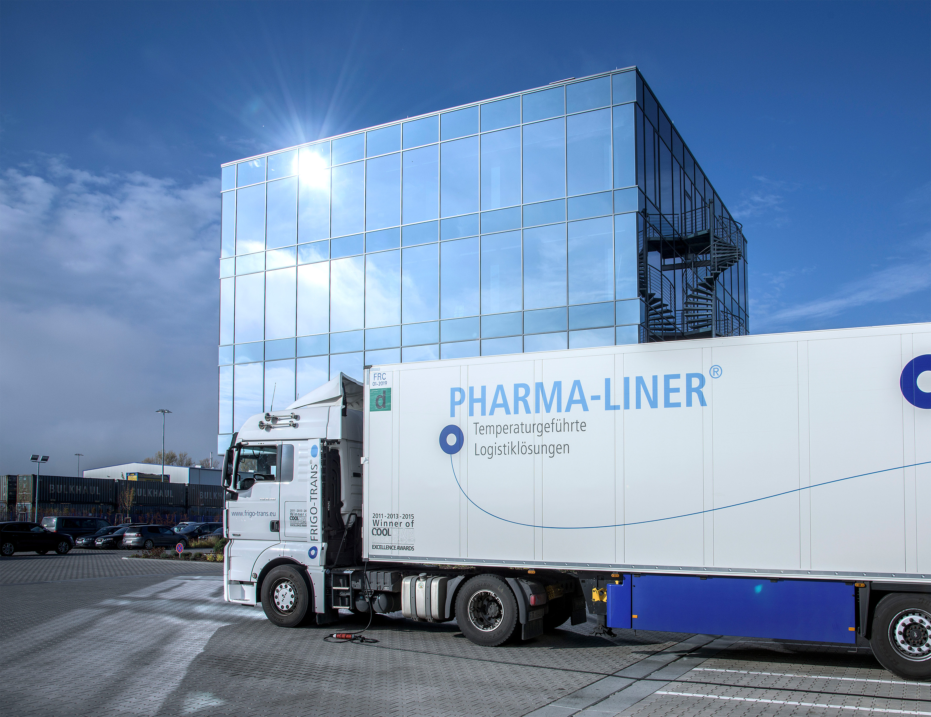 Das Bild zeigt eine Luftaufnahme einer weitläufigen Gebäudeanlage mit roten Ziegeldächern und weißen Fassaden, eingebettet in eine grüne, waldreiche Landschaft. Die Anlage besteht aus mehreren miteinander verbundenen Gebäudeflügeln, die einen rechteckigen Innenhof bilden. Rund um die Gebäude befinden sich Bäume, Wiesen und kleinere Straßen mit geparkten Autos. Im Hintergrund ist ein großer See sichtbar, dessen Ufer von dichtem Wald gesäumt ist. Einzelne kleinere Häuser und Villen liegen verstreut zwischen den Bäumen in Seenähe. Die Szenerie wirkt ruhig, idyllisch und naturnah, mit der Kombination aus historischer Architektur und üppiger Natur.