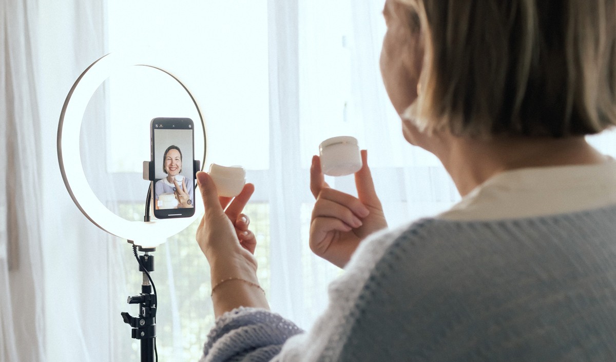 Eine Frau hält einen kleinen Behälter mit Creme in der Hand und spricht in ihr Handy